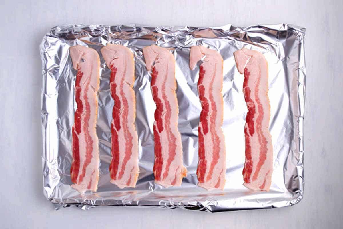 Raw bacon lined up on a foiled-lined baking sheet.