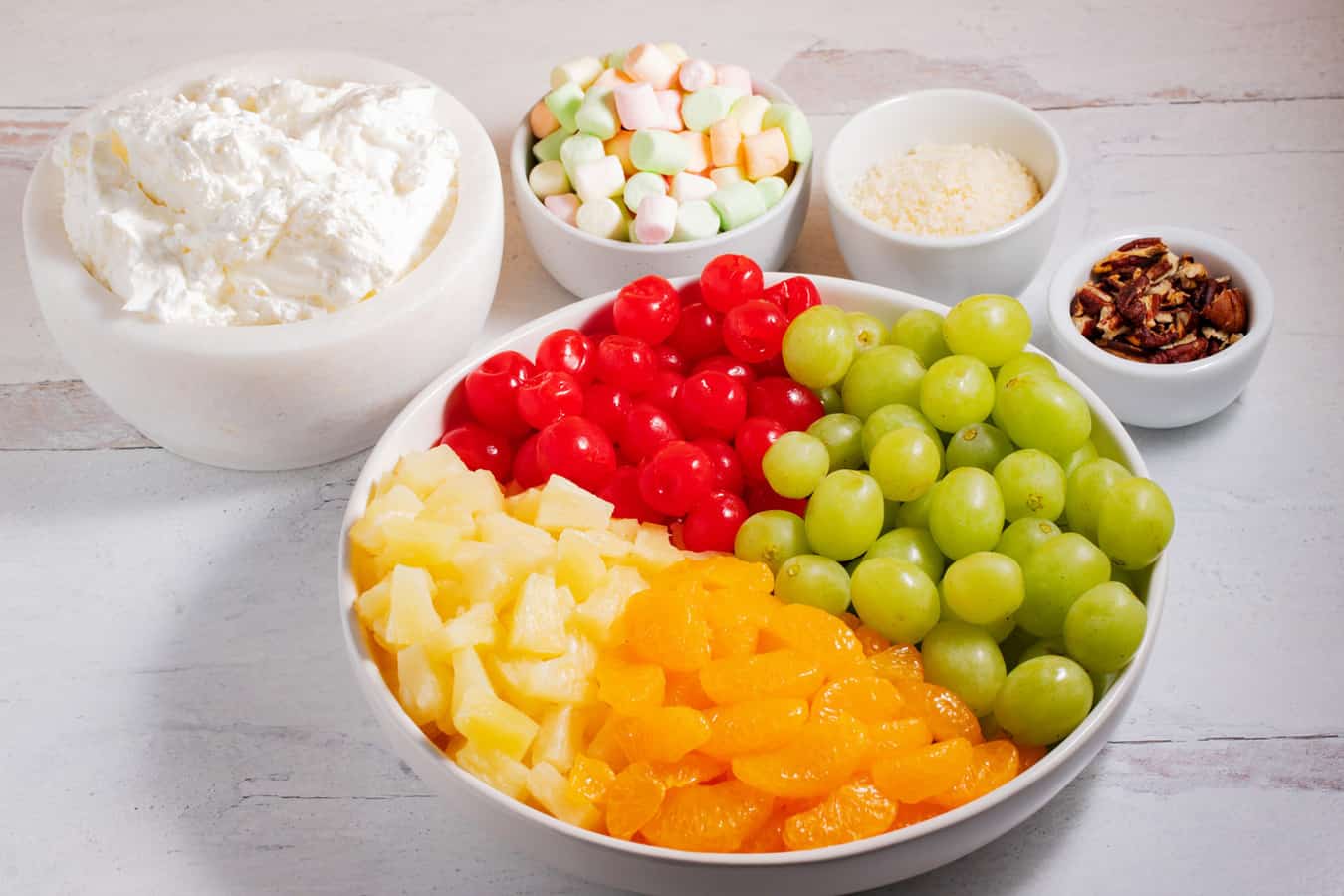 ingredients of ambrosia salad: mandarin oranges, pineapple, grapes, maraschino cherries, creamy sauce, marshmallows, coconut, and pecans