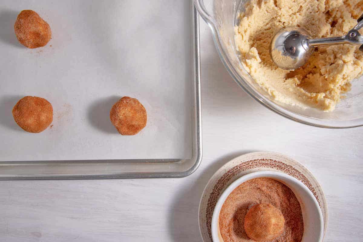 Rolling station with snickerdoodle cookie dough, cinnamon-sugar bowl, and cookie sheet with dough balls on them.