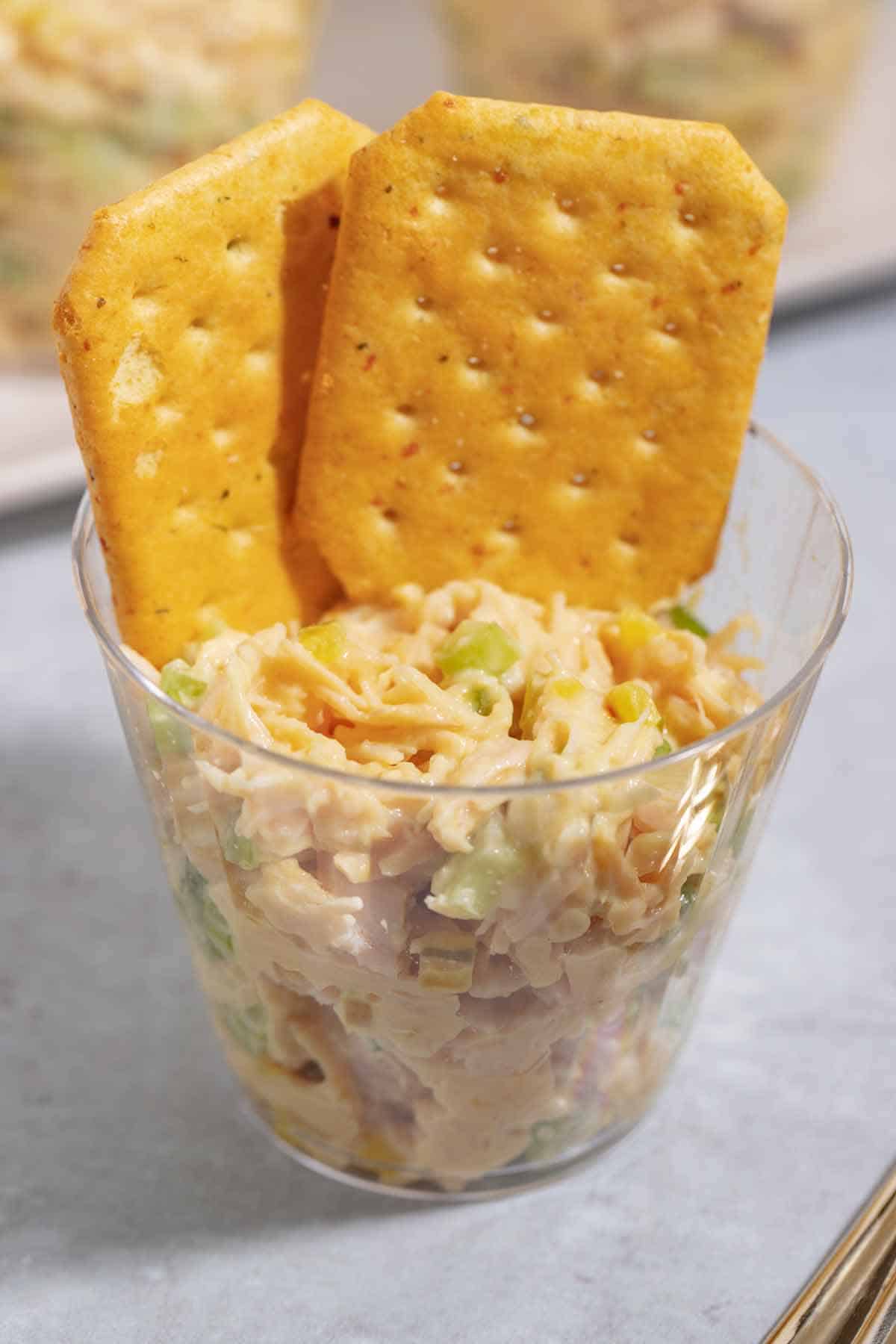 Clear plastic party cup with filled with chicken salad. Two crackers are inside the cup too.
