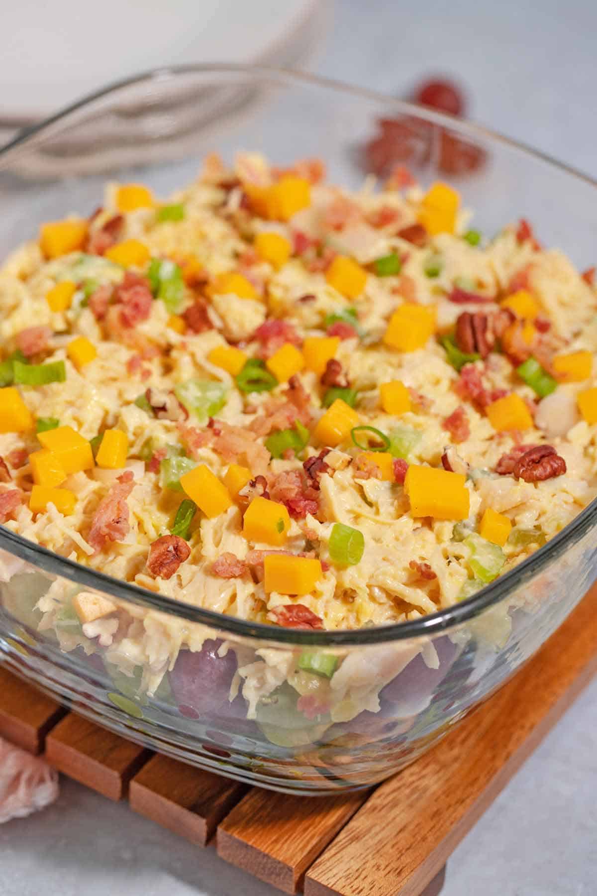 A serving bowl of 7-layer chicken salad. The layers from bottom to top are shredded lettuce, red and green grapes, chicken salad, green onions, cheddar cheese cubes, pecan pieces, and crumbled bacon.