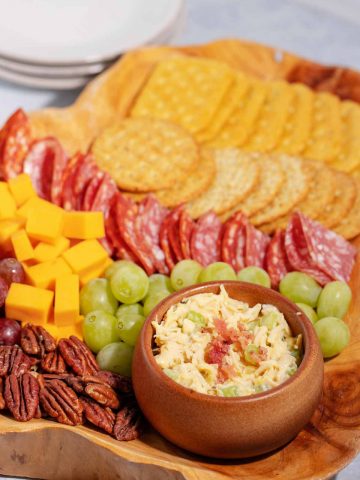 A charcuterie board with a bowl of chicken salad surrounded by pecans, cheese cubes, red and green grapes, salame, and two rows of crackers.