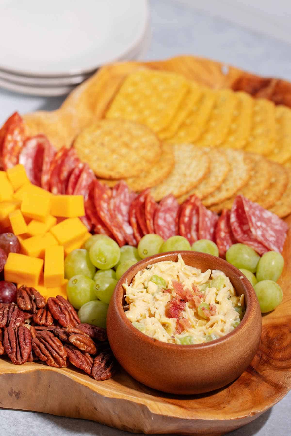 A charcuterie board with a bowl of chicken salad surrounded by pecans, cheese cubes, red and green grapes, salame, and two rows of crackers.