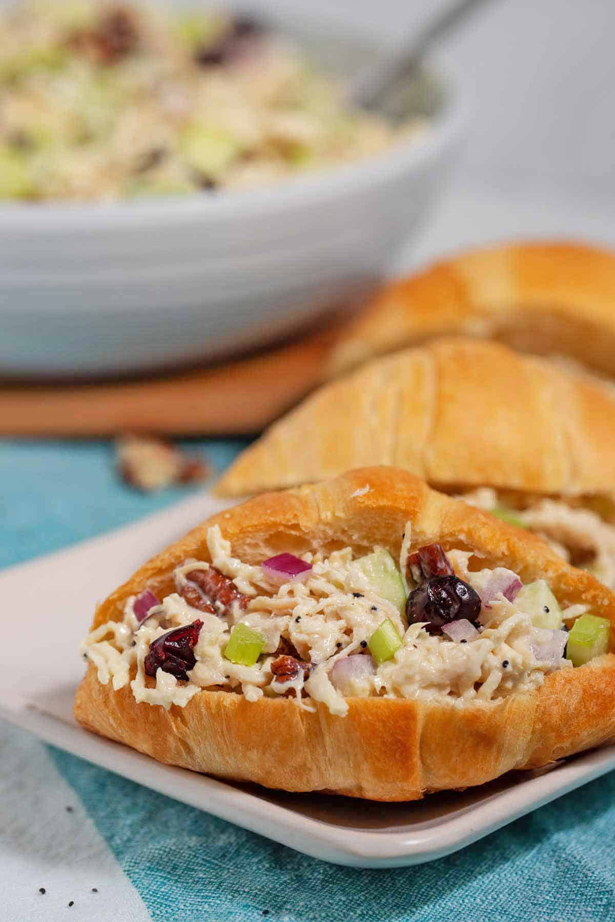 Chicken salad croissant sandwiches on a plate with a bowl of chicken salad in the background.