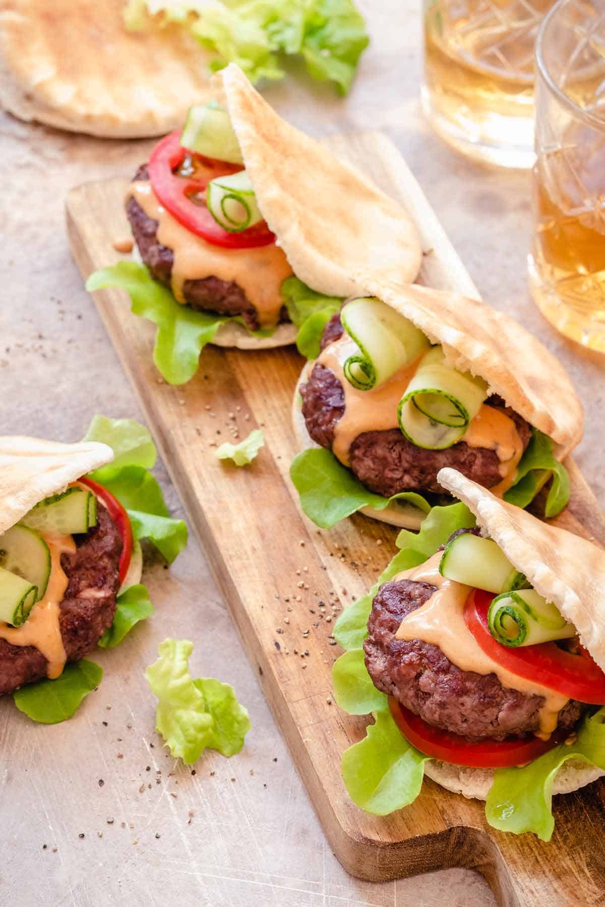 Sandwich board with 3 pita burgers. Lettuce, tomato, sauce, shaved cucumber, and burger are stuffed inside pita bread.