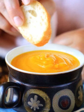 A hand dipping crusty bread into a bowl of pumpkin soup.