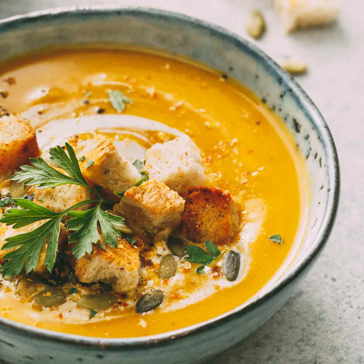 Bowl of pumpkin soup garnished with cream, croutons, parsley, and pepitas.