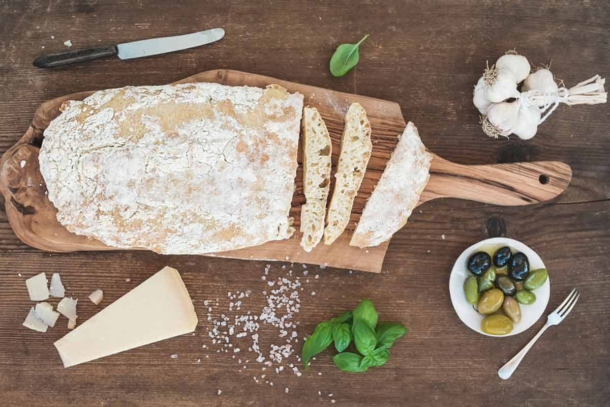 Loaf of crusty bread on cutting board surrounded by Parmesan cheese, olives, basil, and garlic.