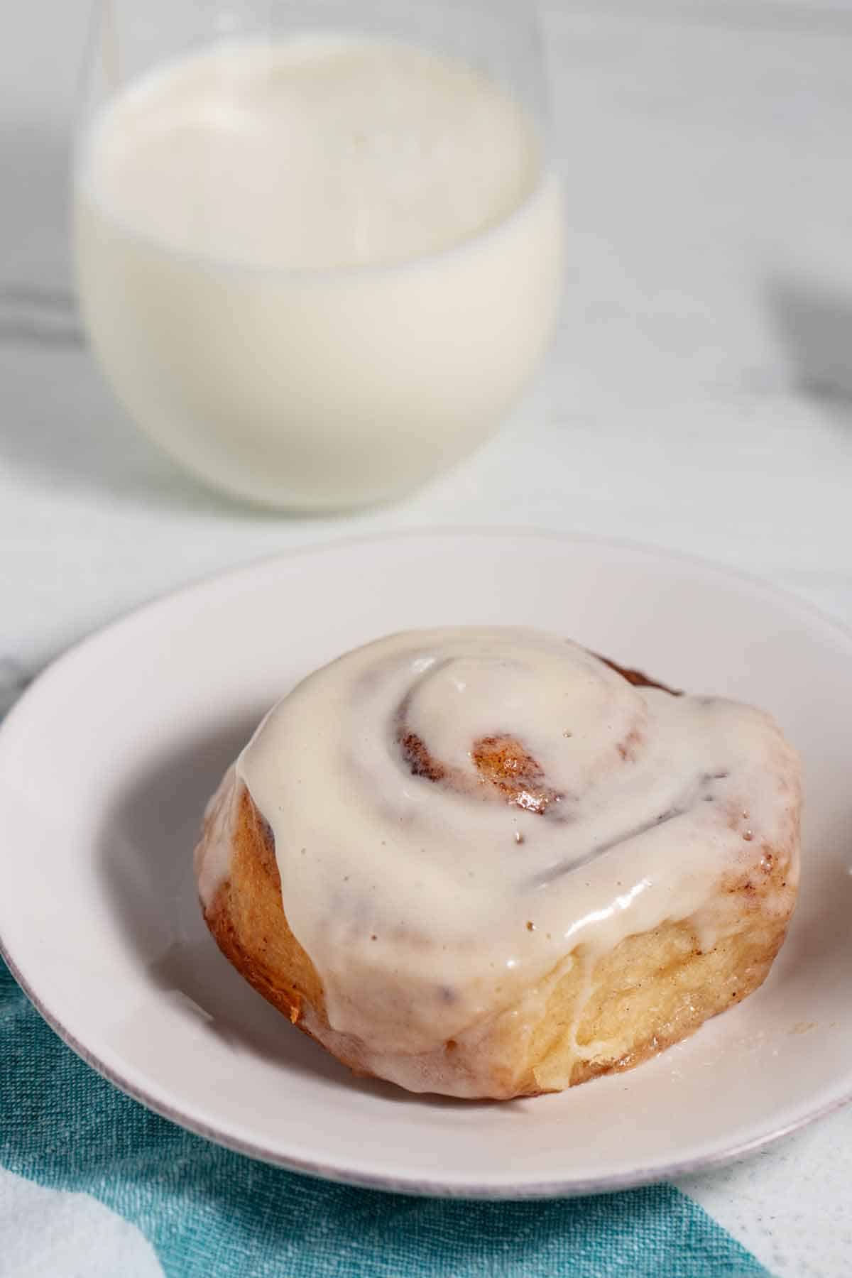 Cinnamon roll with icing on a plate and glass of milk in the background.