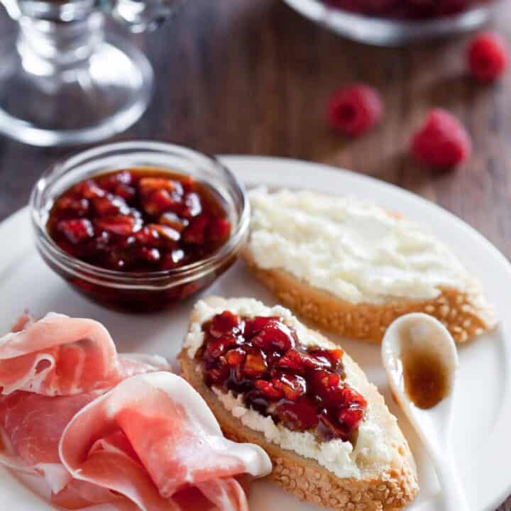 Plate of crostini smeared with ricotta cheese and topped with cranberries with prosciutto on the side.