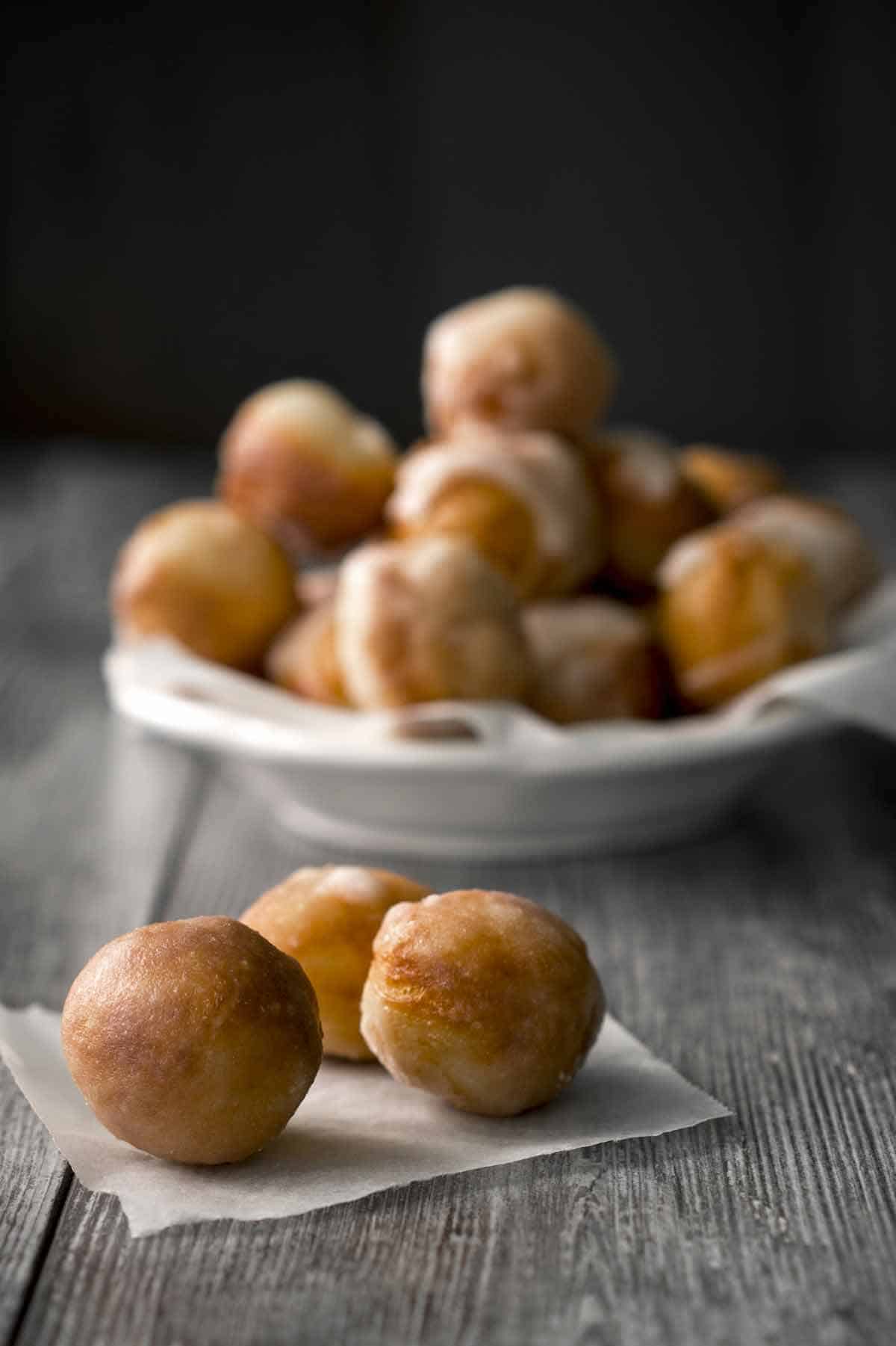 Serving bowl of donut holes with 3 donut holes on a parchment sheet in the foreground.