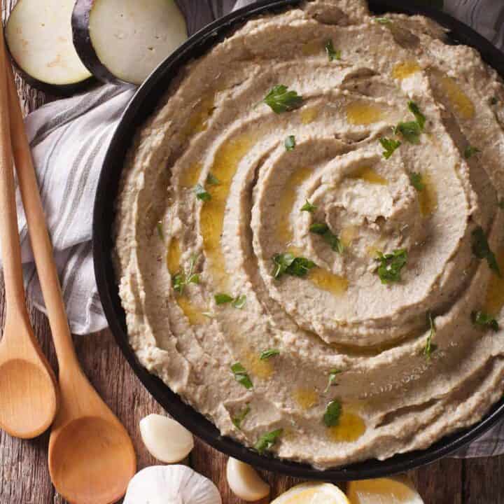 Bowl of eggplant dip garnished with olive oil and parsley. The bowl is surrounded by garlic cloves, sliced eggplant, and lemon.