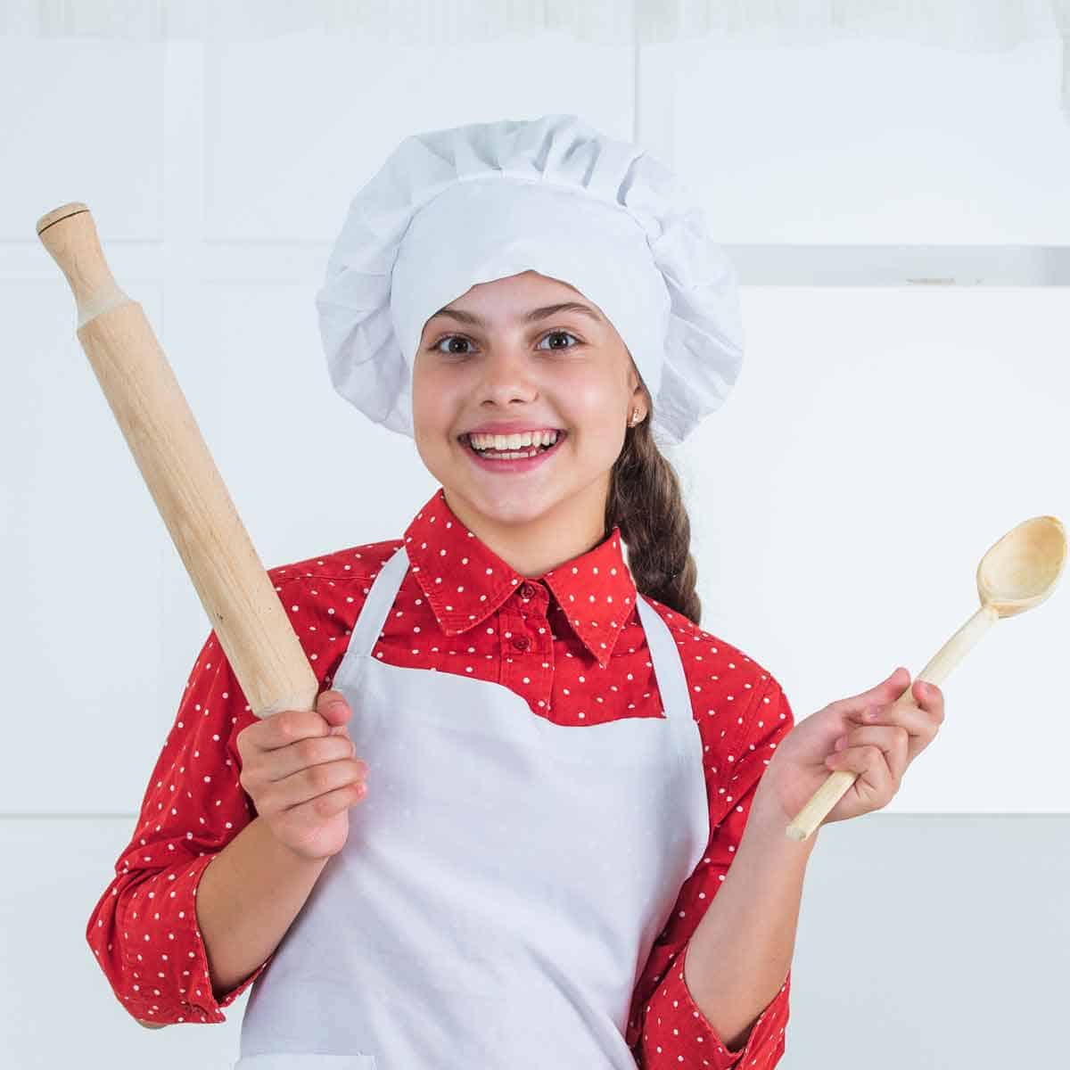 Tween girl with chef hat and apron holding a wooden spoon and rolling pin.