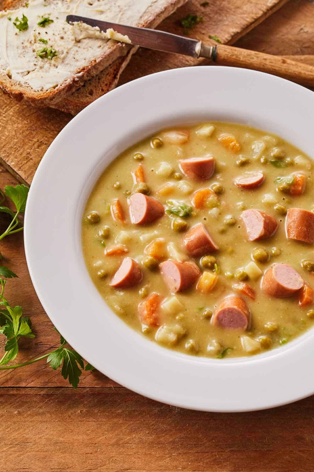 Bowl of split pea soup with sausage and a side of bread and butter on a plate.