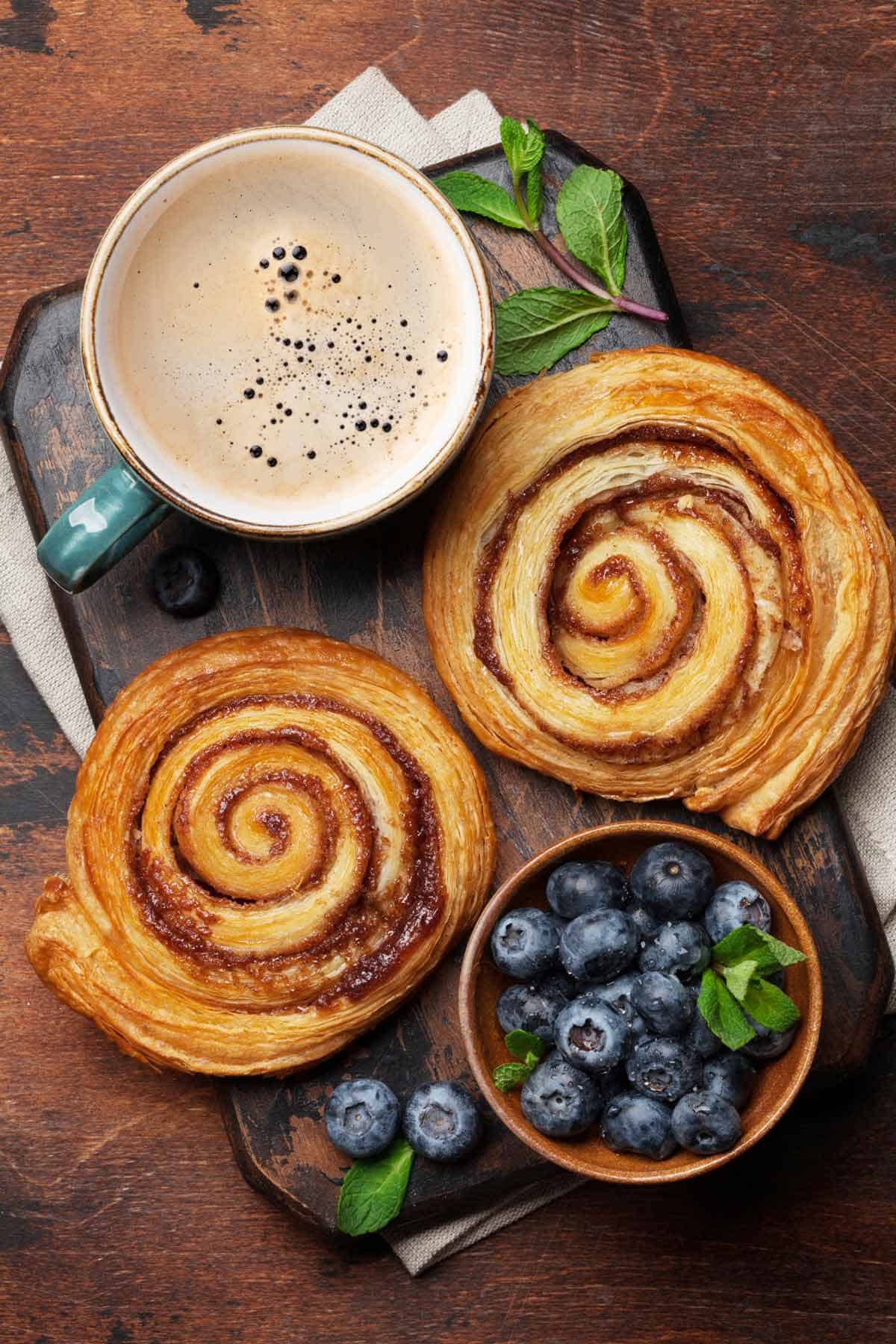 Two cinnamon rolls on a bread board with a bowl of blueberries and a cup of hot chocolate.