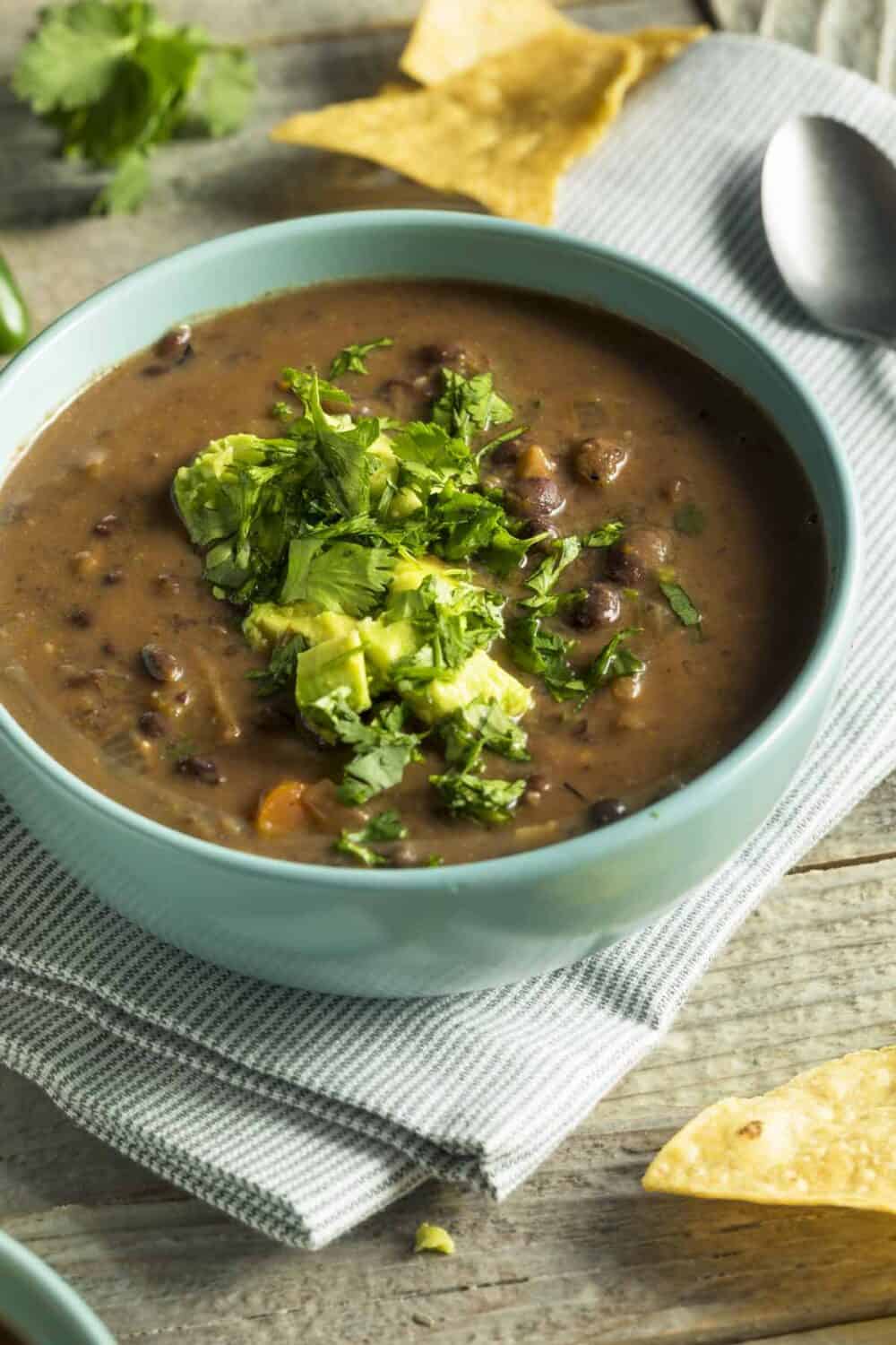 Bowl of black bean soup garnished with cilantro and avocado chunks. Tortilla chips are in the background.