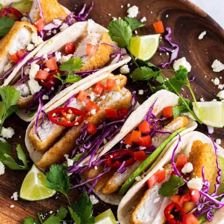 Serving bowl with fish tacos. They are surrounded by lime quarters, fresh cilantro, and Cotija cheese crumbles.