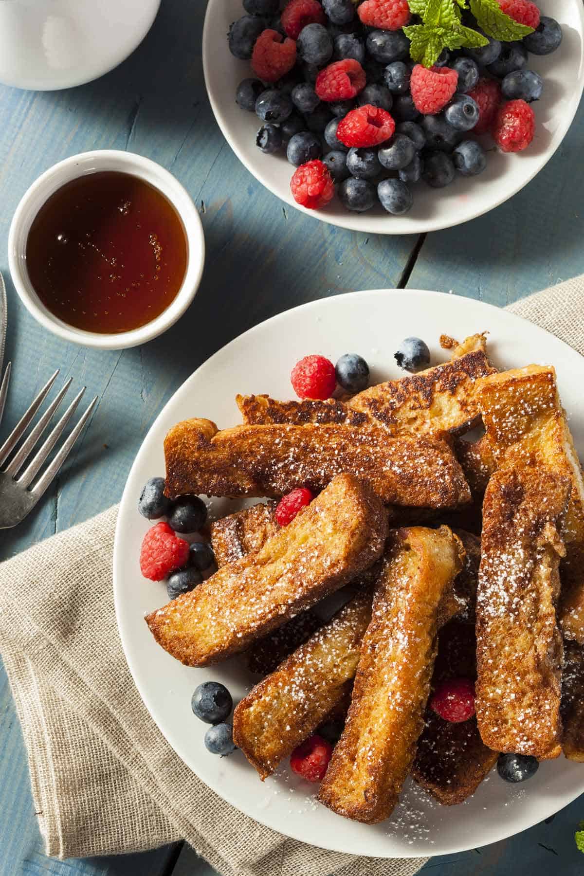 Plate of French toast sticks garnished wtih fresh berries and powdered sugar. Plate of berry fruit salad and small bowl of syrup is in the background.