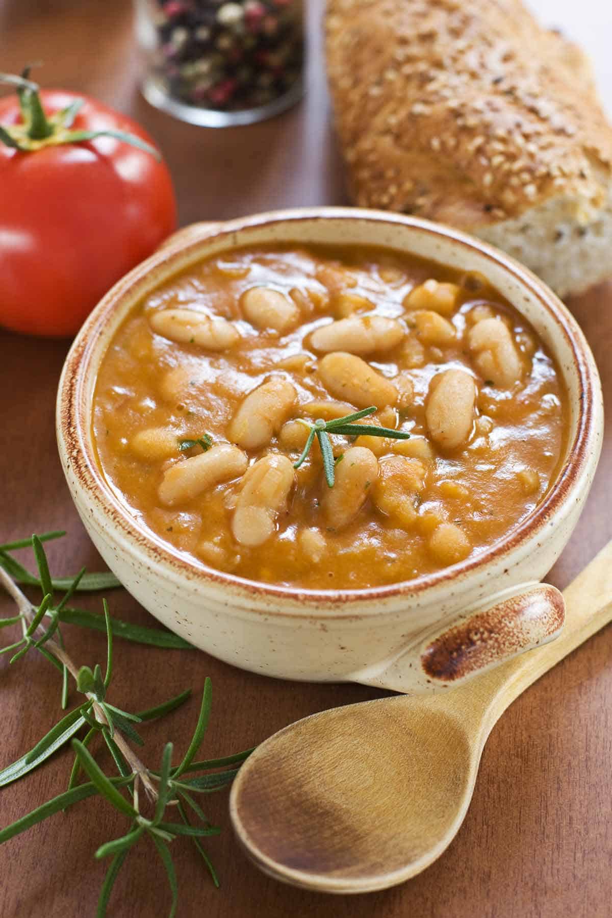 Bowl of navy bean soup garnished with rosemary. A tomato, wooden spoon, and crusty bread are in the background.