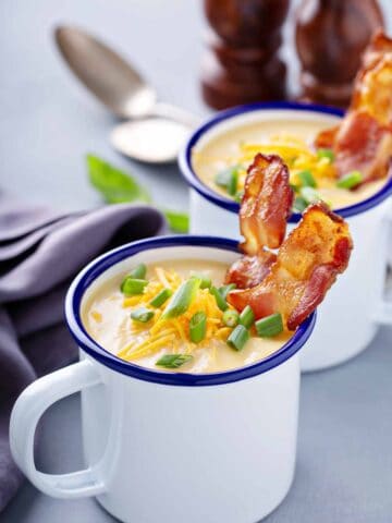 Two mugs of potato soup garnished with shredded cheese, sliced green onions, and strips of bacon.