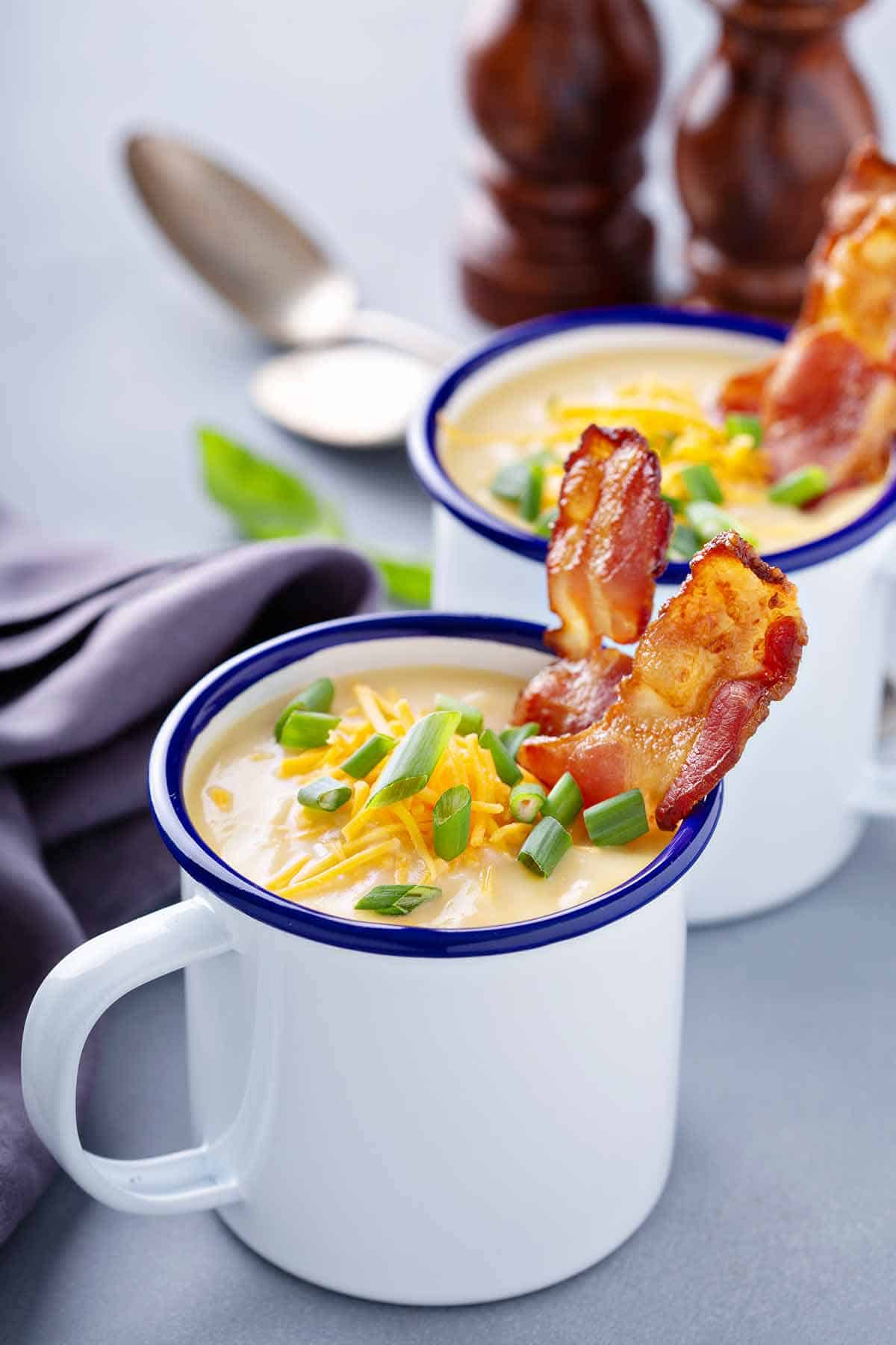 Two mugs of potato soup garnished with shredded cheese, sliced green onions, and strips of bacon.