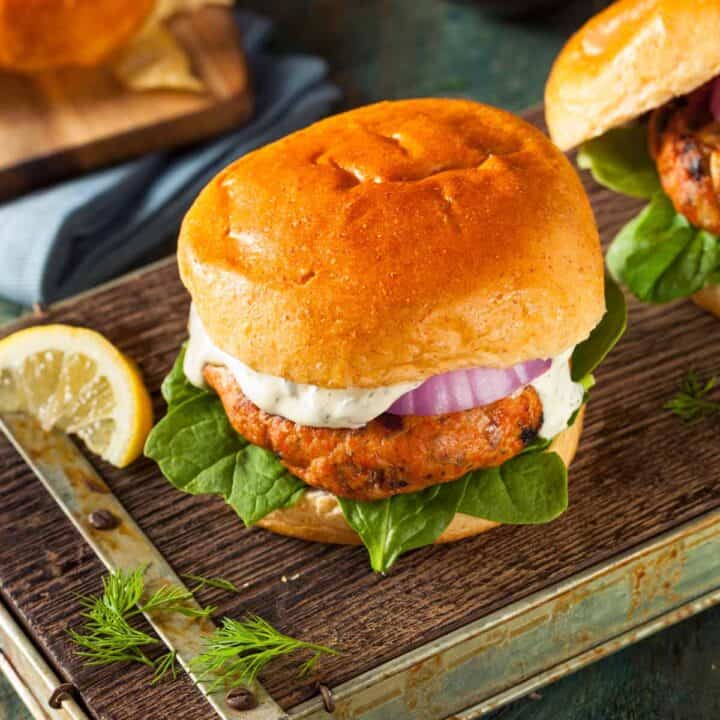 Salmon burger on a serving board. It has tartar sauce, sliced red onion and leafy greens.