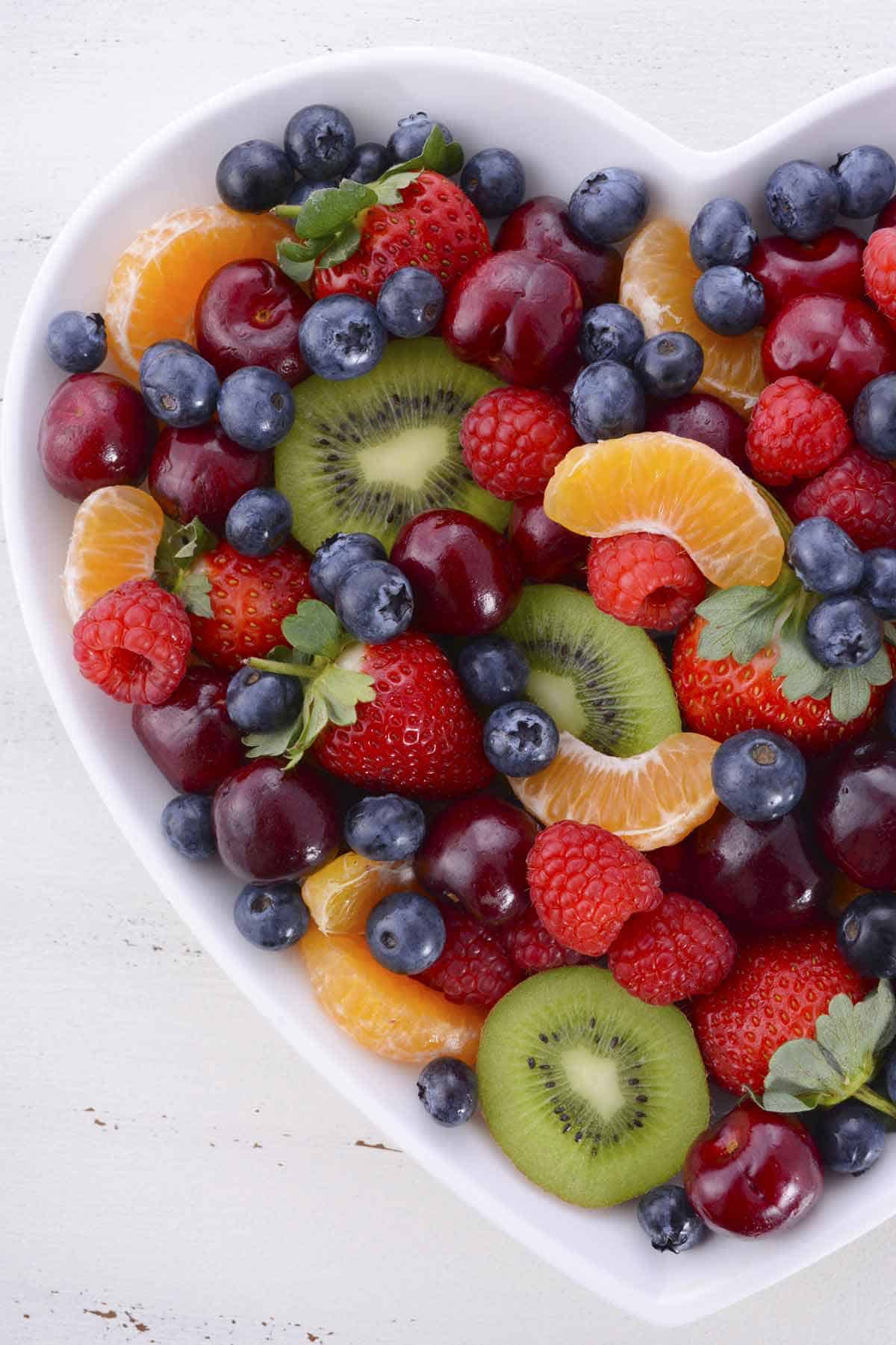 Heart shaped bowl filled with fruit salad. Salad has strawberries, kiwi, raspberries, blueberries, cherries, and orange wedges.