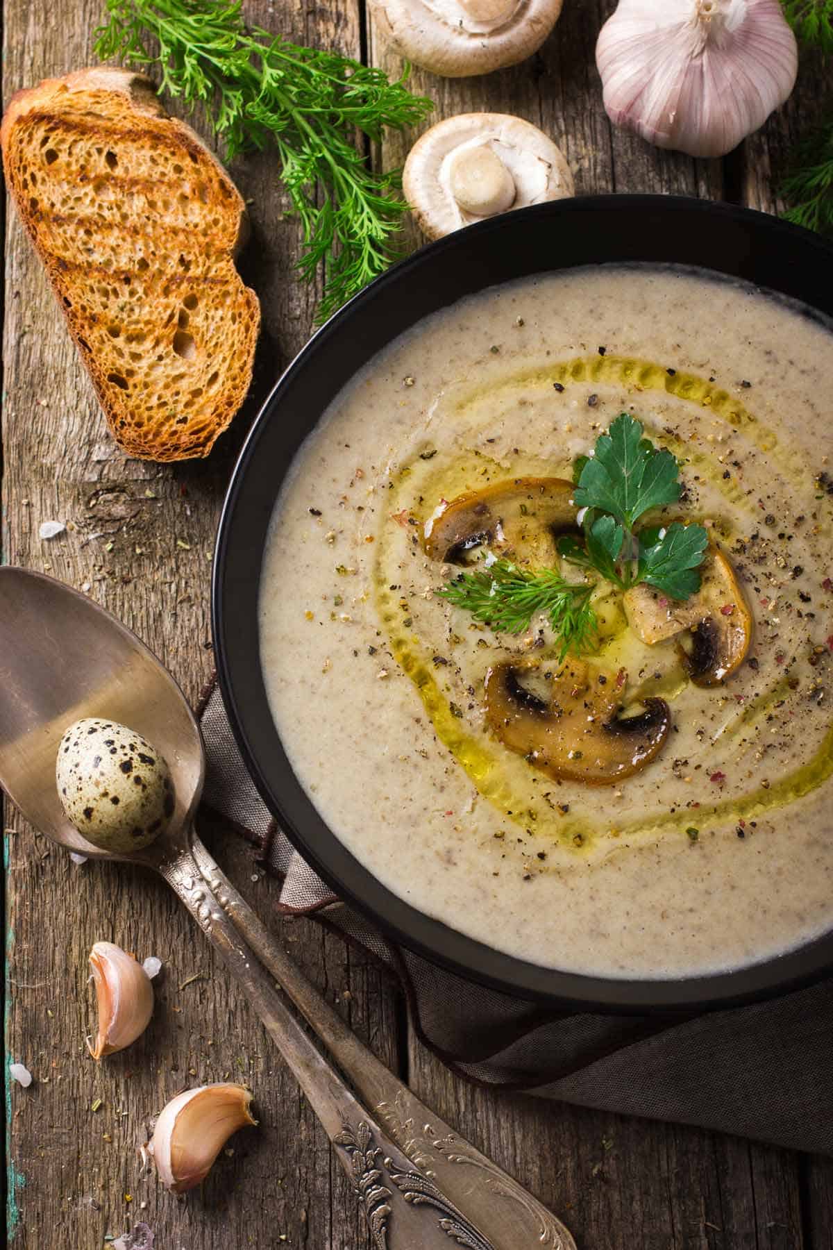 Bowl of creamy mushroom soup with toast, mushrooms, and garlic in the background.