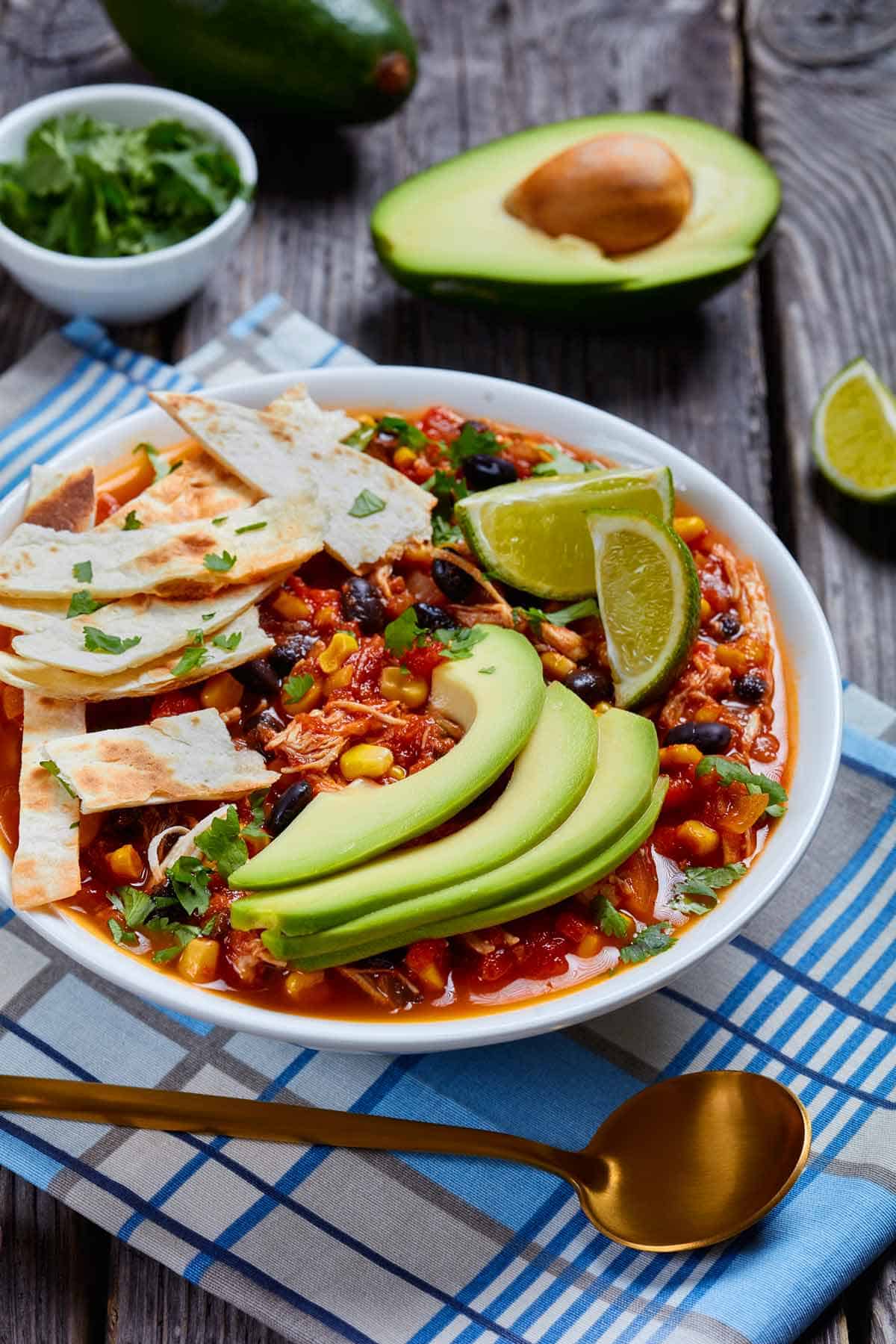 Bowl of taco soup with tortilla strips, sliced avocado, and lime wedges.