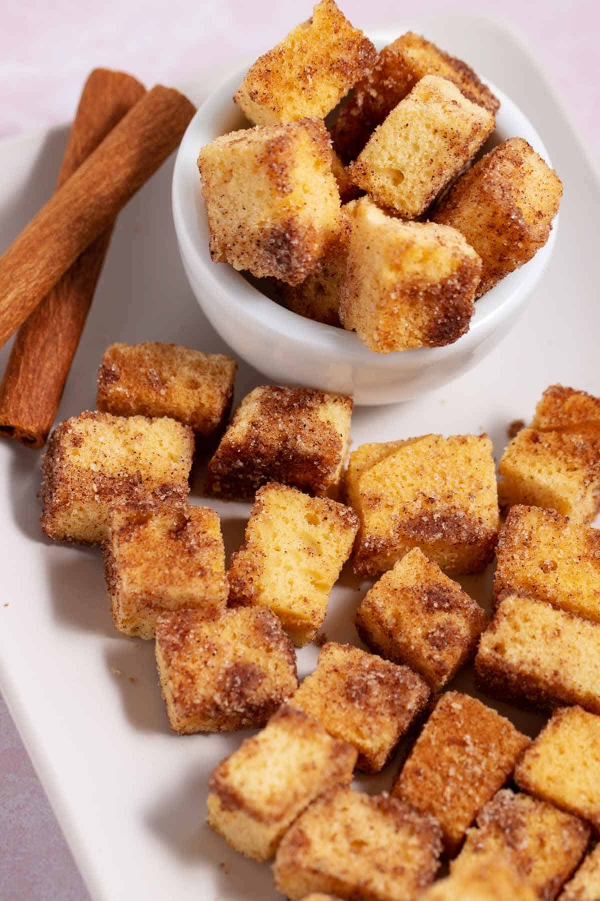 Bowl and plate filled with cinnamon sugar pound cake croutons. Two cinnamon sticks are on the plate too.