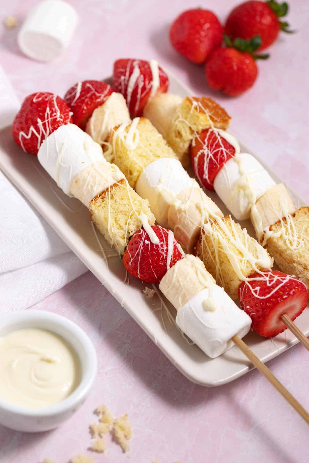 Long dessert skewers with strawberries, bananas, big marshmallows, and cake cubes drizzled with white chocolate with whole strawberries in the background.