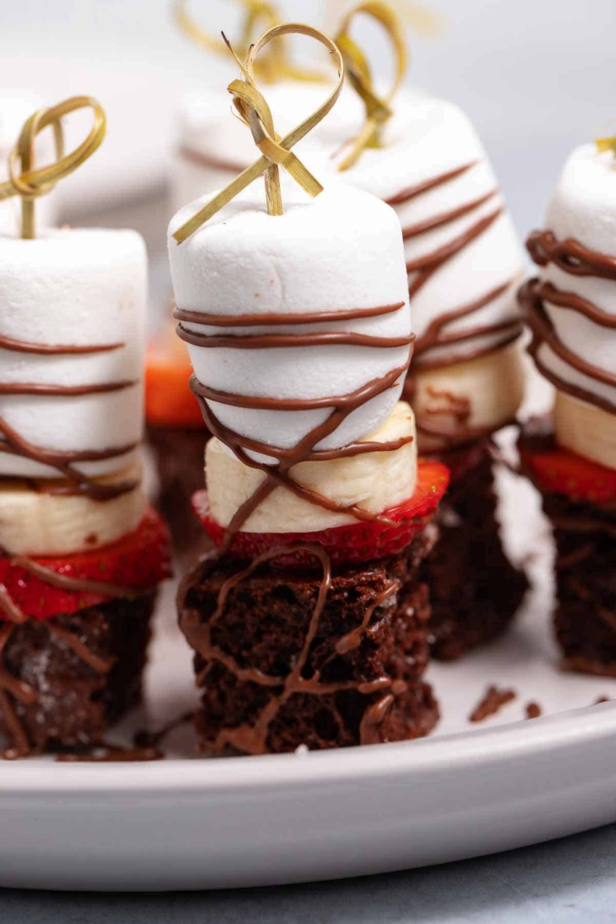 Plate of dessert skewers with a giant marshmallow, banana, strawberry, and chocolate cake croutons.
