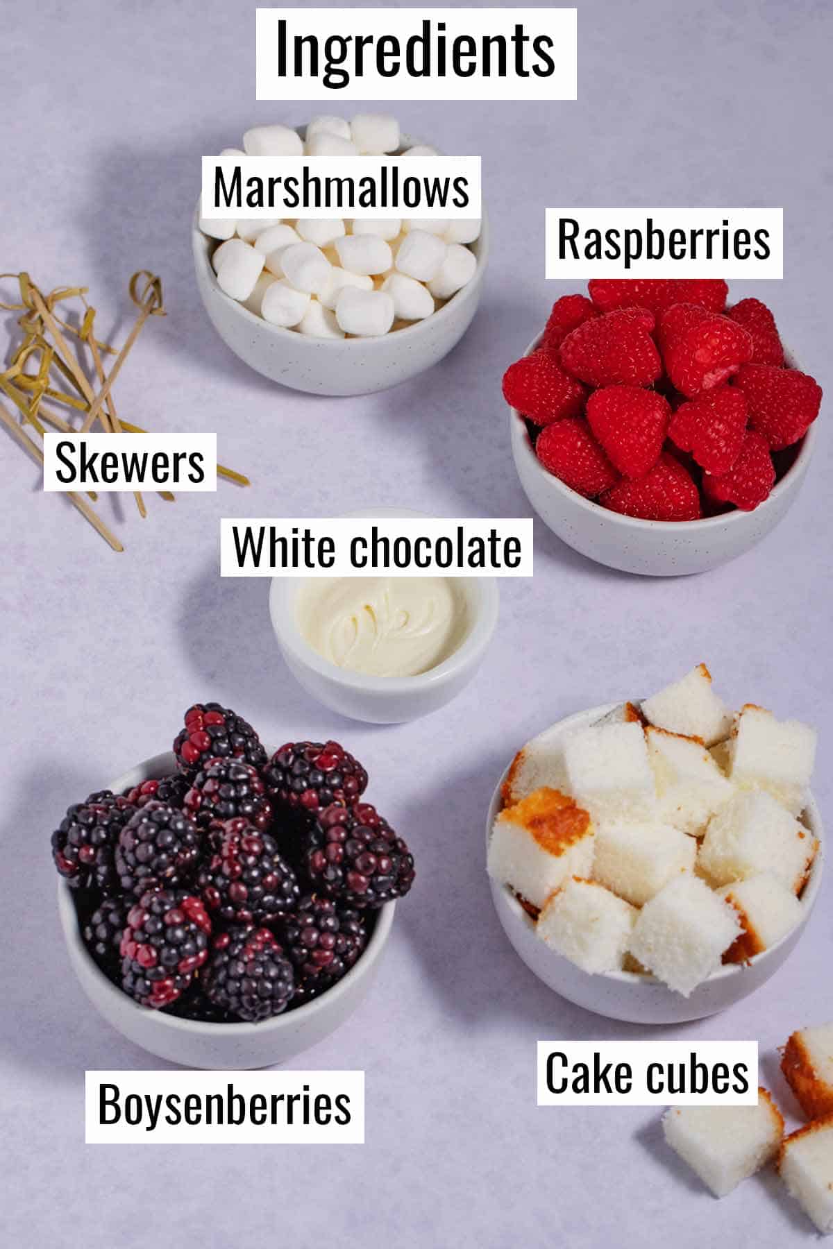 Ingredients for dessert skewers. Bowls of raspberries, boysenberries, cake cubes, marshmallows, and melted white chocolate.