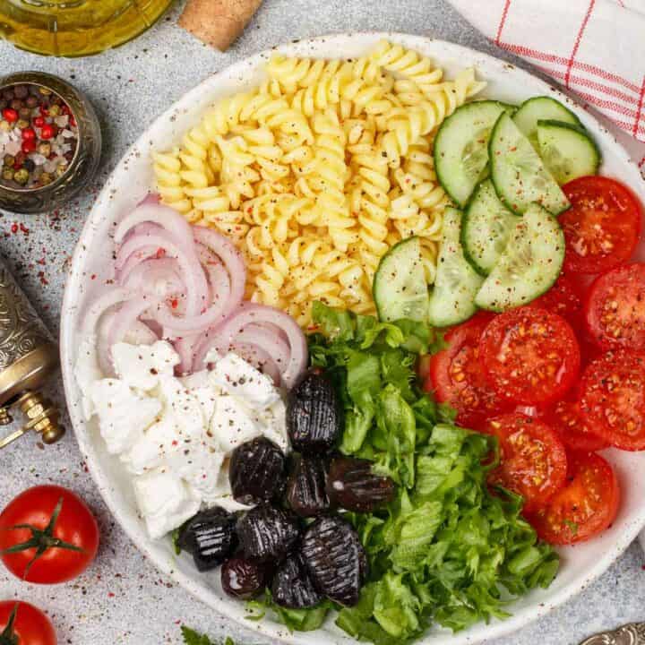 Greek pasta salad on a plate with noodles, cucumbers, tomatoes, olives, feta cheese, and red onions.