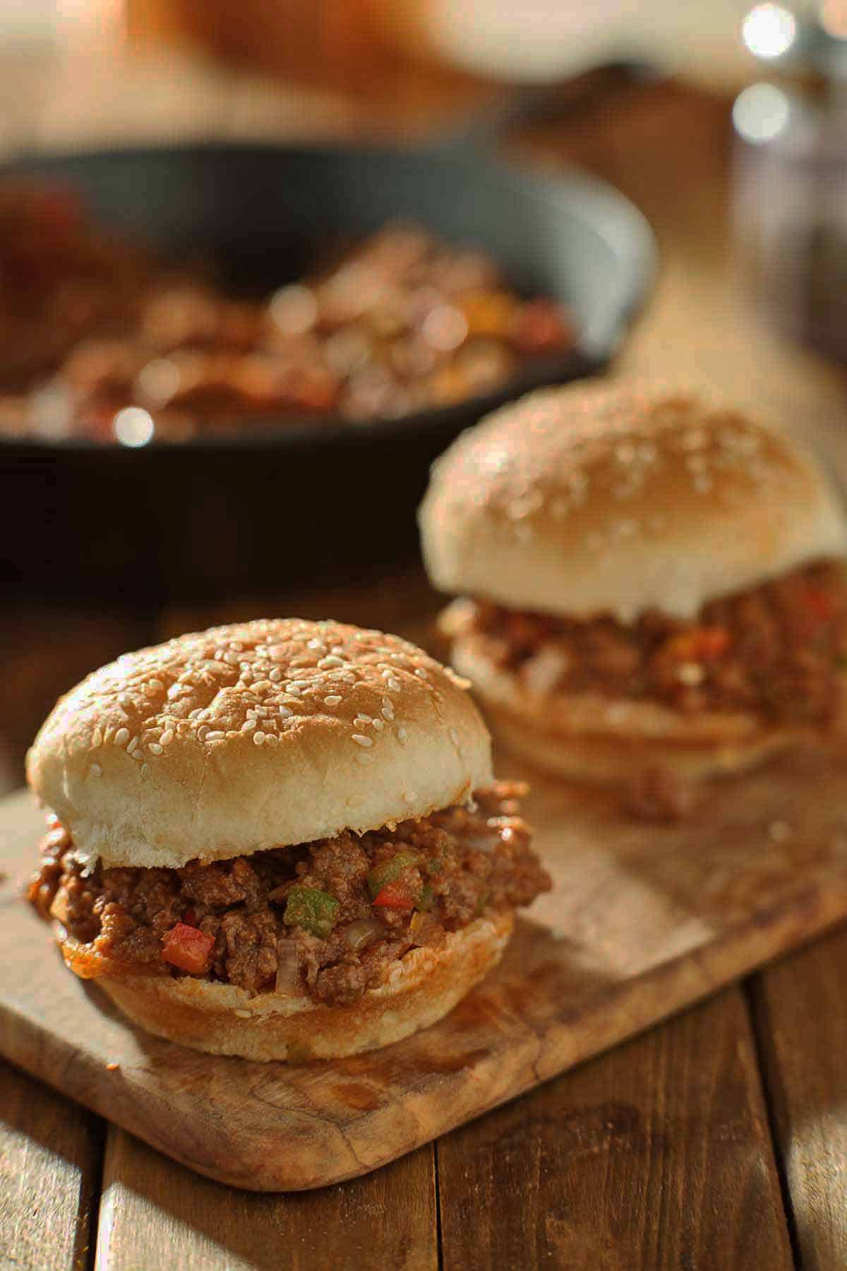 Two sloppy joes on a sandwich board with leftover filling in a pan.