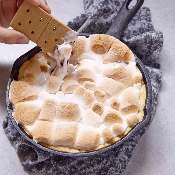 Smores dip in a hot skillet with a hand dipping a graham cracker.