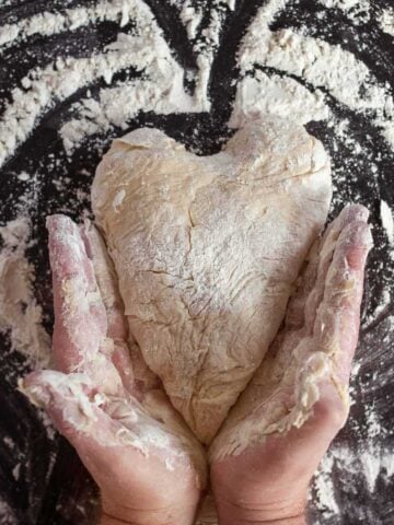 Pizza dough on a floured surface shaped into a heart with two hands holding it on the outside.