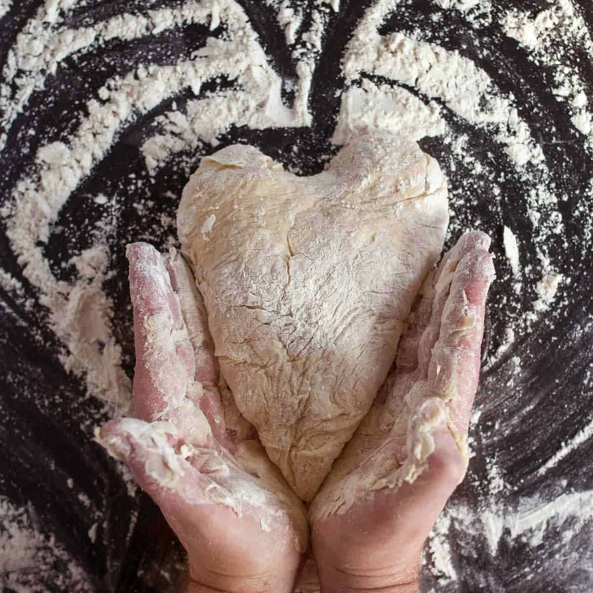 Pizza dough on a floured surface shaped into a heart with two hands holding it on the outside.