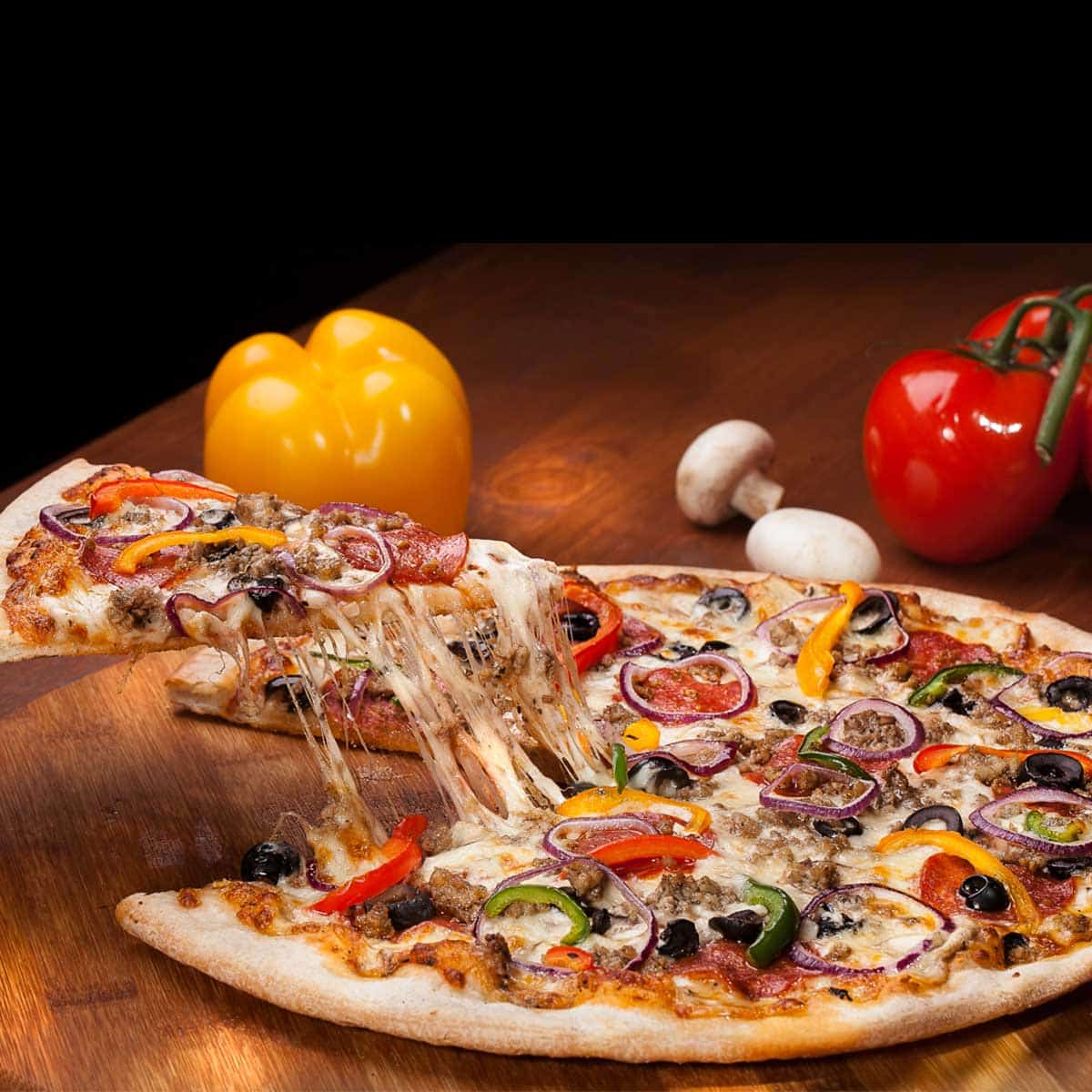 A whole loaded topping pizza on a platter with one slice being pulled out showing the strings of cheese.