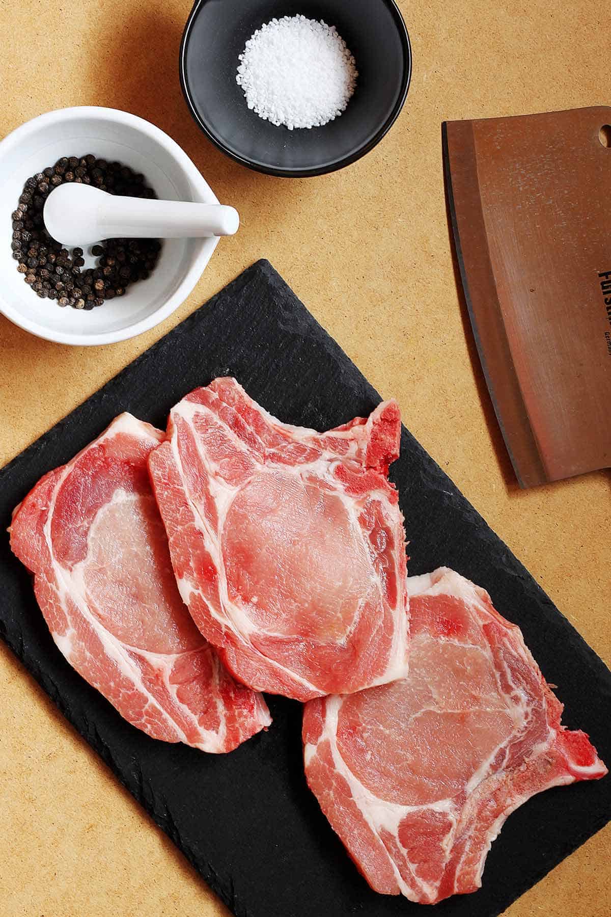 Raw meat on a cutting board with pepper corns and kosher salt in separate bowls nearby.