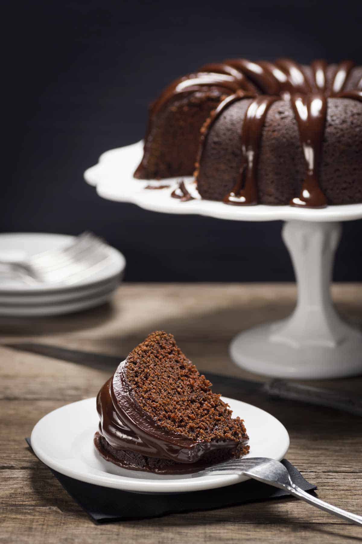 Slice of chocolate Bundt cake decorated with ganache on a plate. The full cake is on a cake stand in the background.