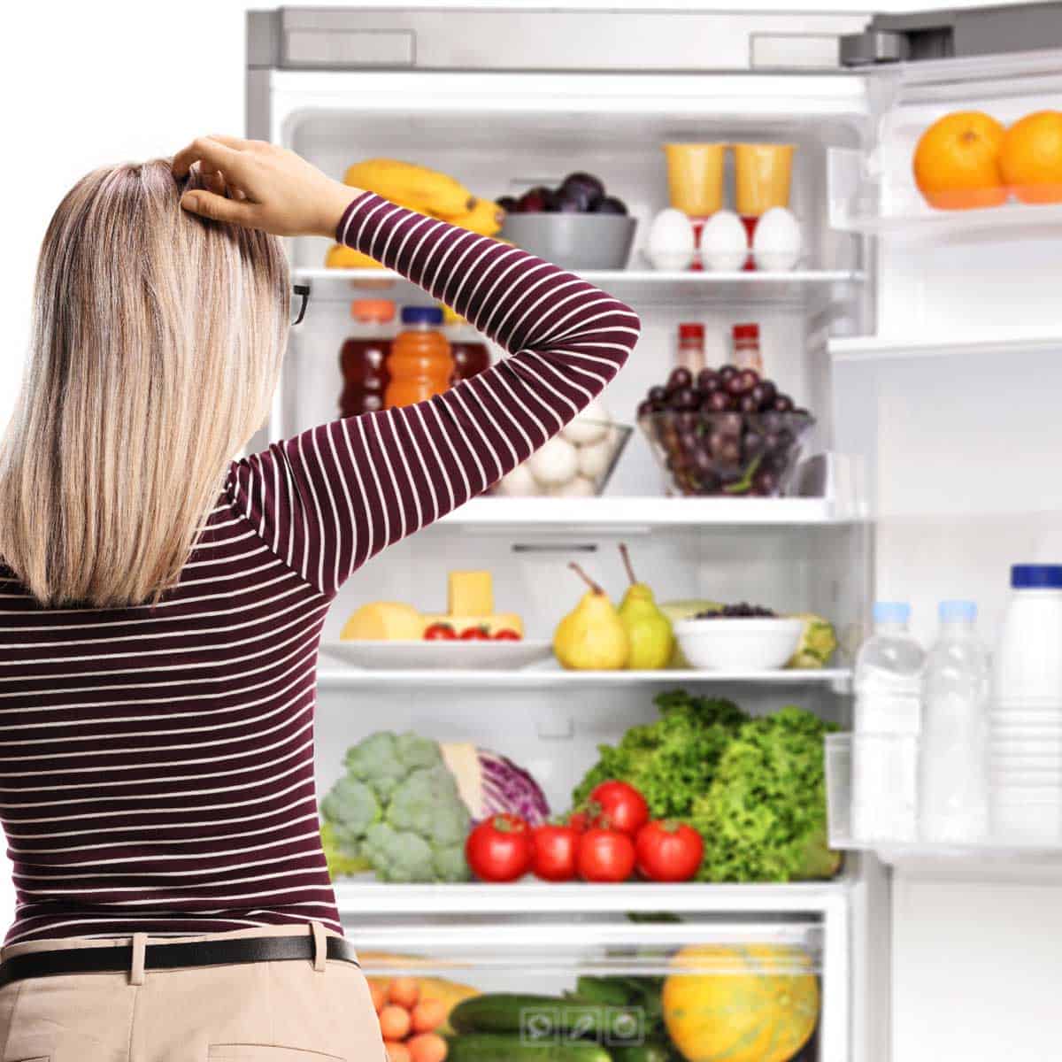 Woman standing at the open door of the fridge looking at the items on each shelf.