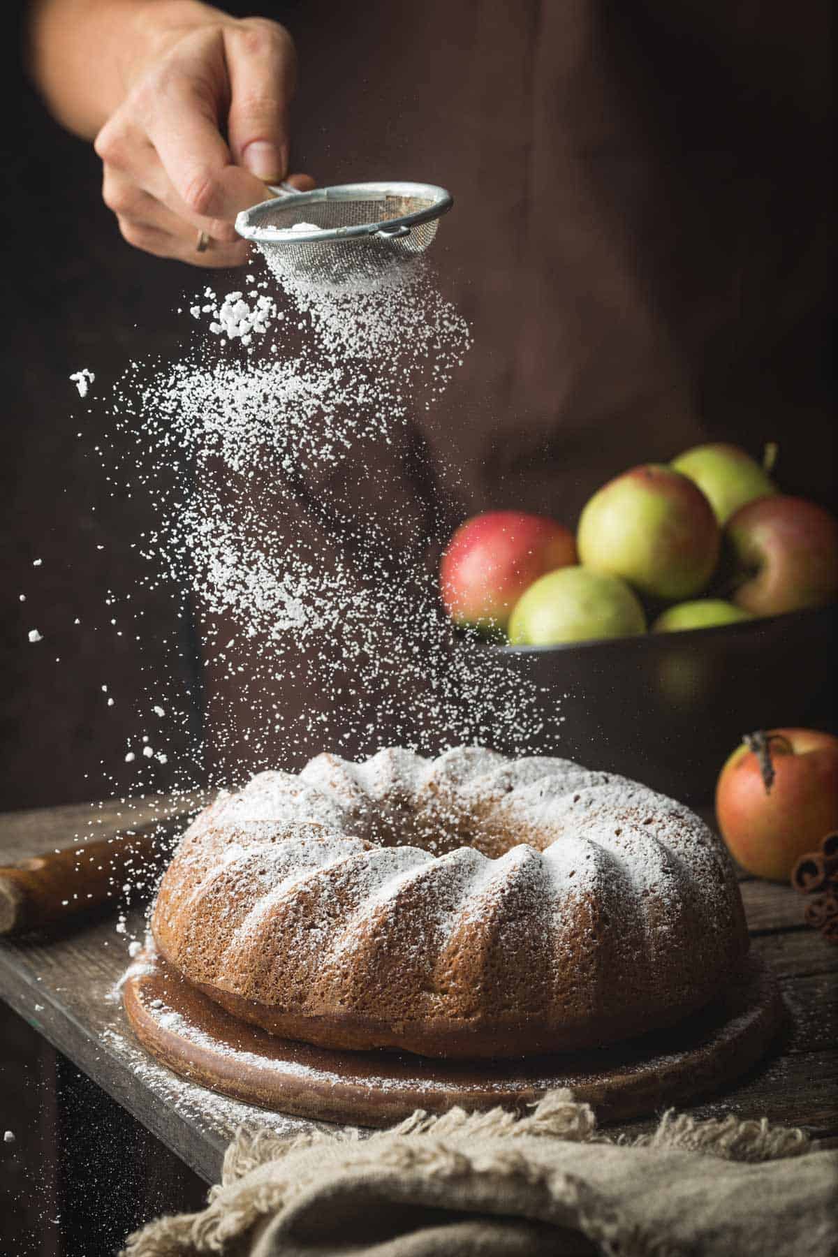 Dusting powdered sugar on a Bundt cake with a box of apples in the background.