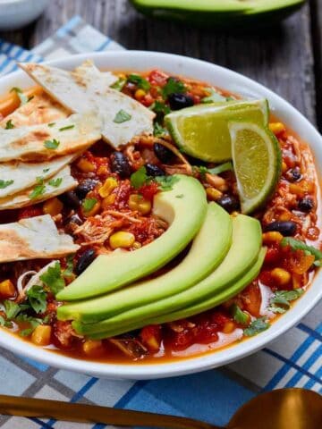 Bowl of taco soup garnished with tortilla strips, avocado slices, and lime wedges.