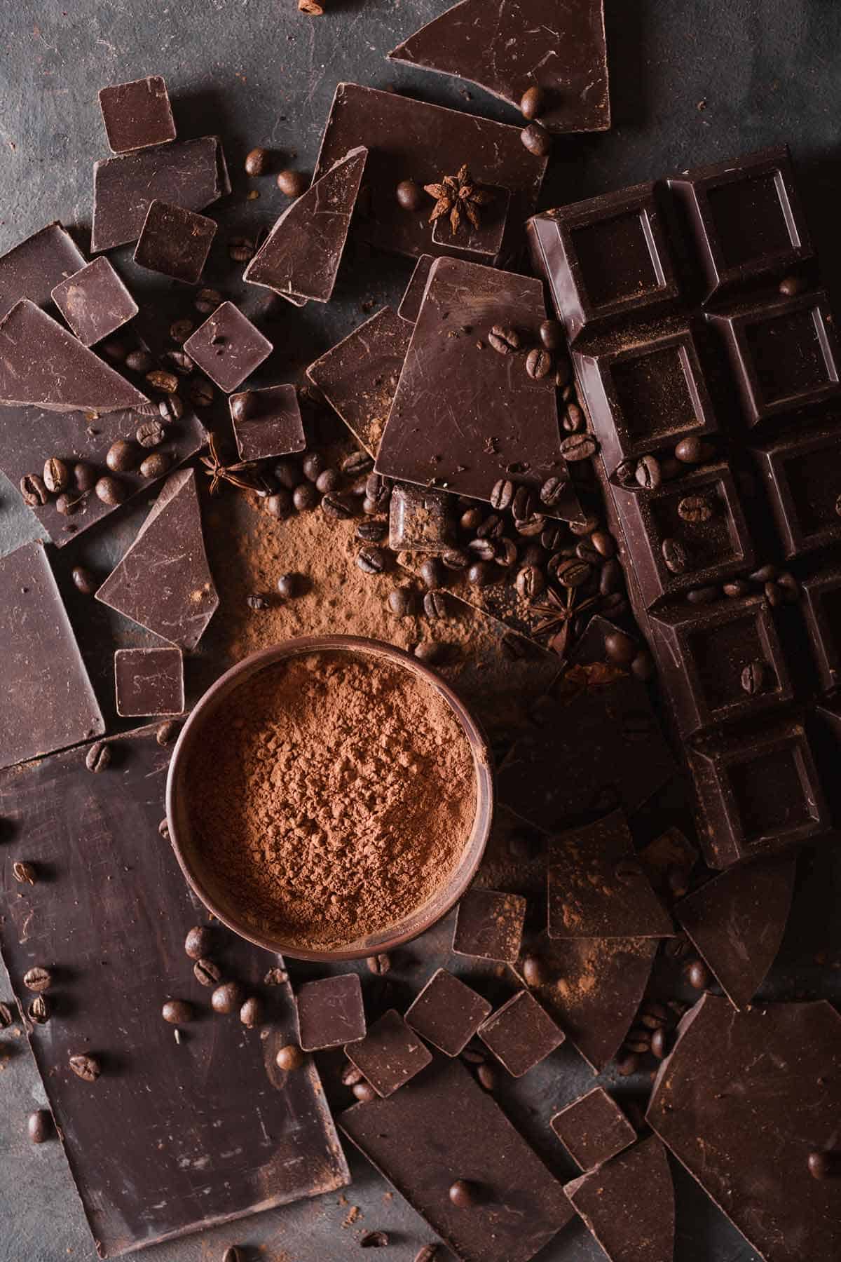 A bowl of cocoa powder surrounded by unsweetened bars of chocolate and cocoa beans.