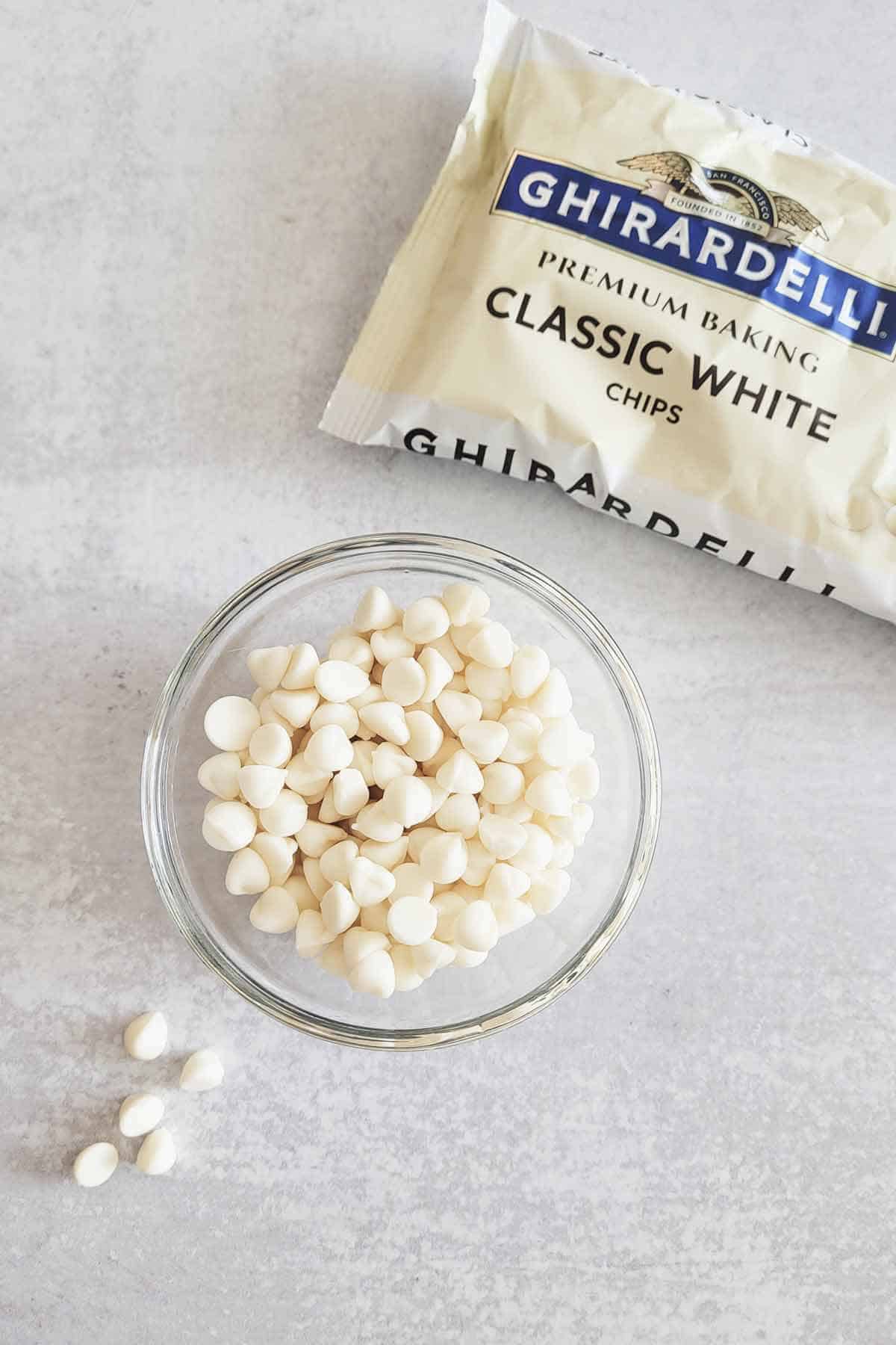 Package of Ghirardelli classic white chips in the background and a bowl of them in the foreground.