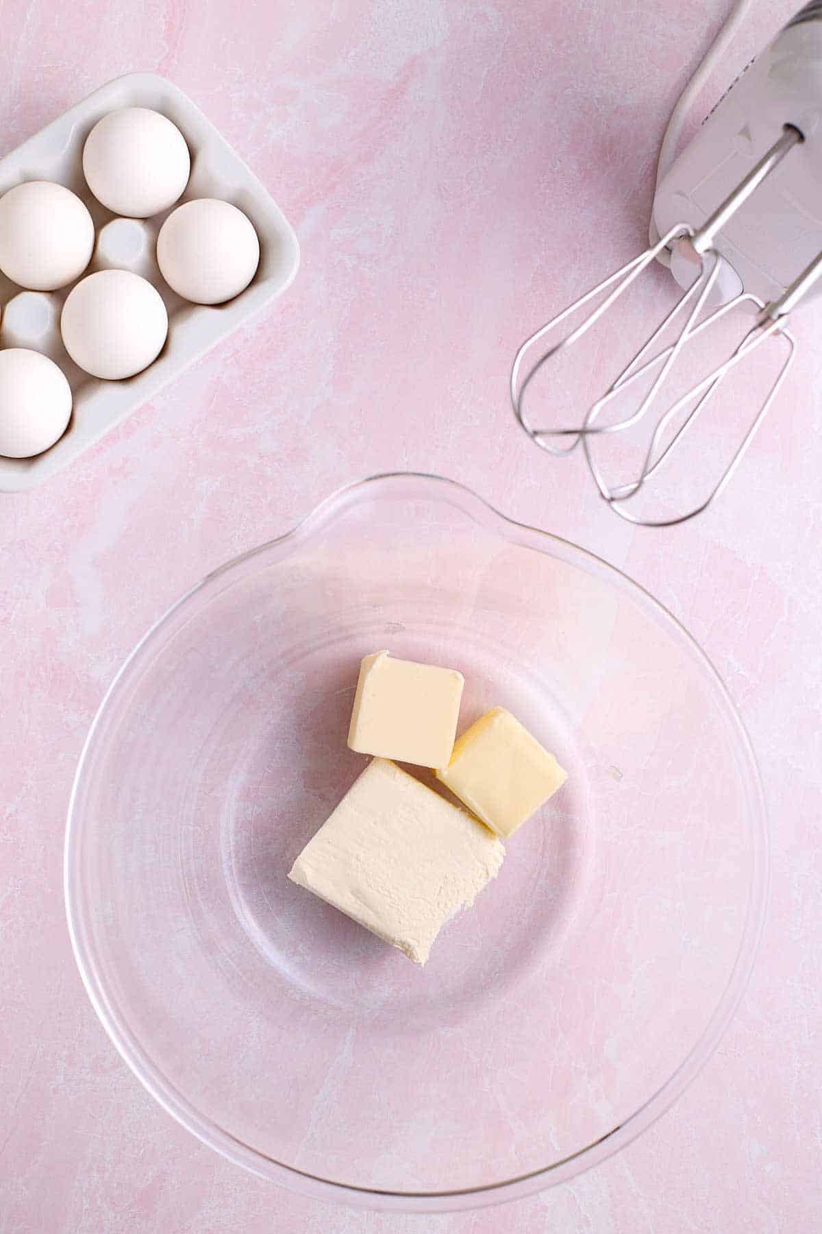 Mixing bowl with softened butter and cream cheese. Electric mixer and eggs in the background.