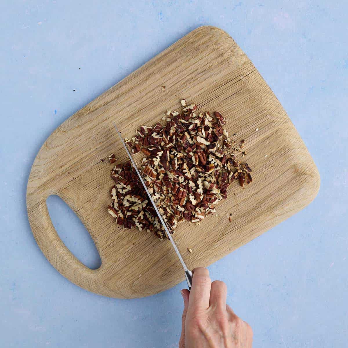Chopping toasted pecans with a knife on a cutting board.