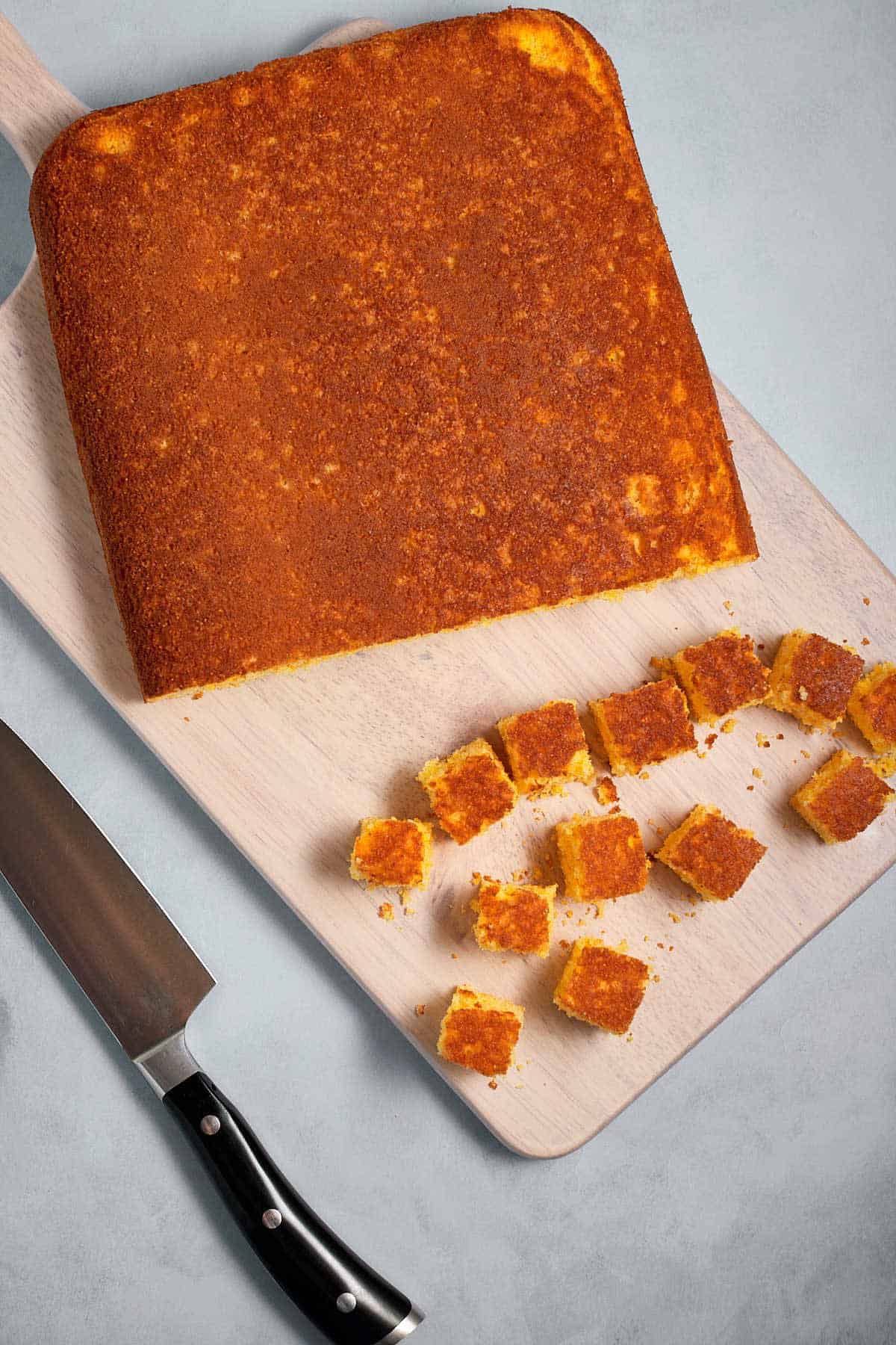 Baked cornbread being cut into cubes on a cutting board.