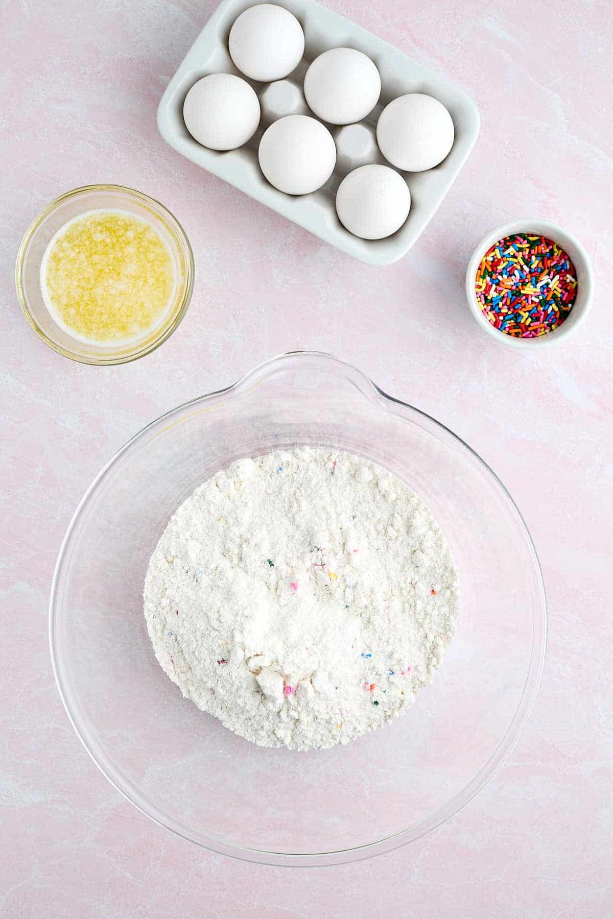 Mixing bowl with Funfetti cake mix and vanilla pudding mix with melted butter, eggs, and sprinkles in bowls surrounding it.
